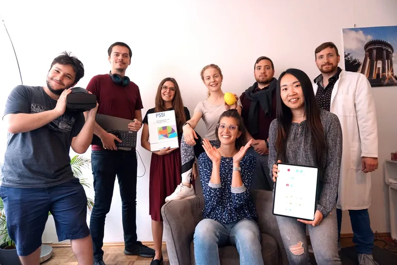 Teamfoto von brainboost: acht Teammitglieder mit Gegenständen aus ihren Berufsfeldern