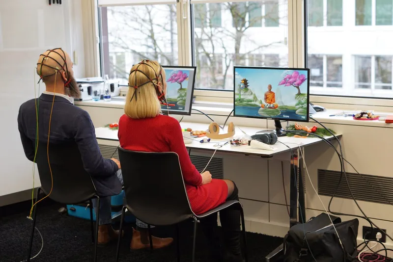 Two people wearing EEG caps during neurofeedback training in front of screens showing a meditating monk